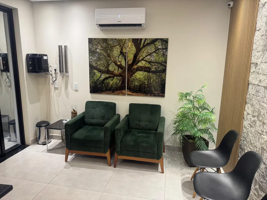 Sala de espera com duas poltronas verdes, quadro de uma árvore na parede, planta e cadeiras pretas.