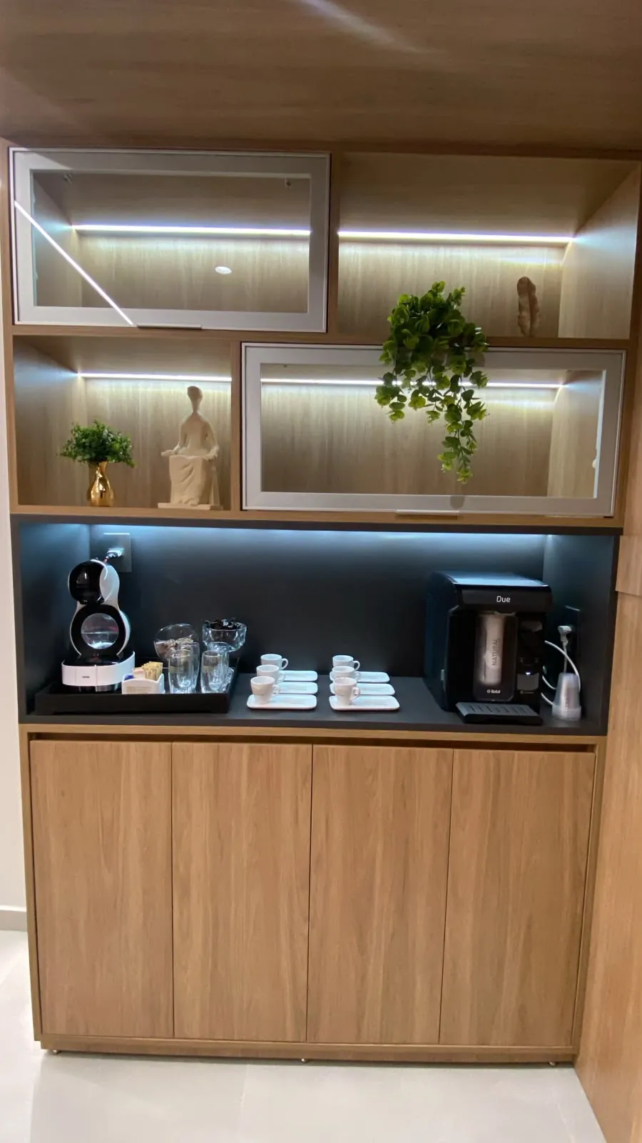 Espaço do café com armário de madeira iluminado, máquina de café, xícaras e purificador de água.