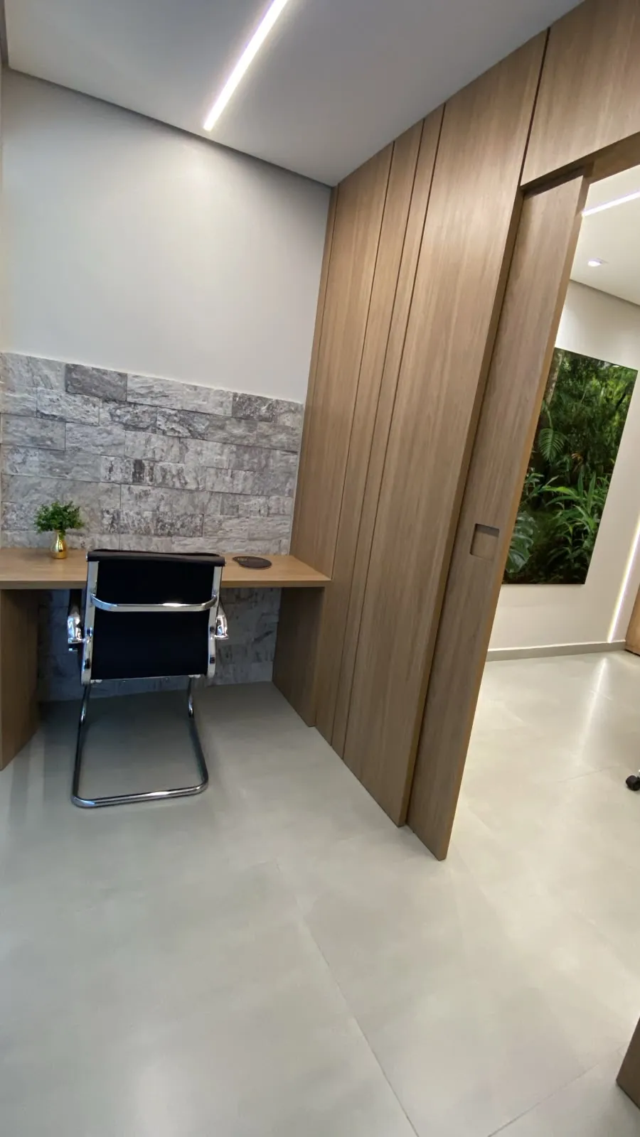 Circulação com a estação de trabalho à esquerda e a passagem para a sala de reunião, com um quadro de vegetação na parede.