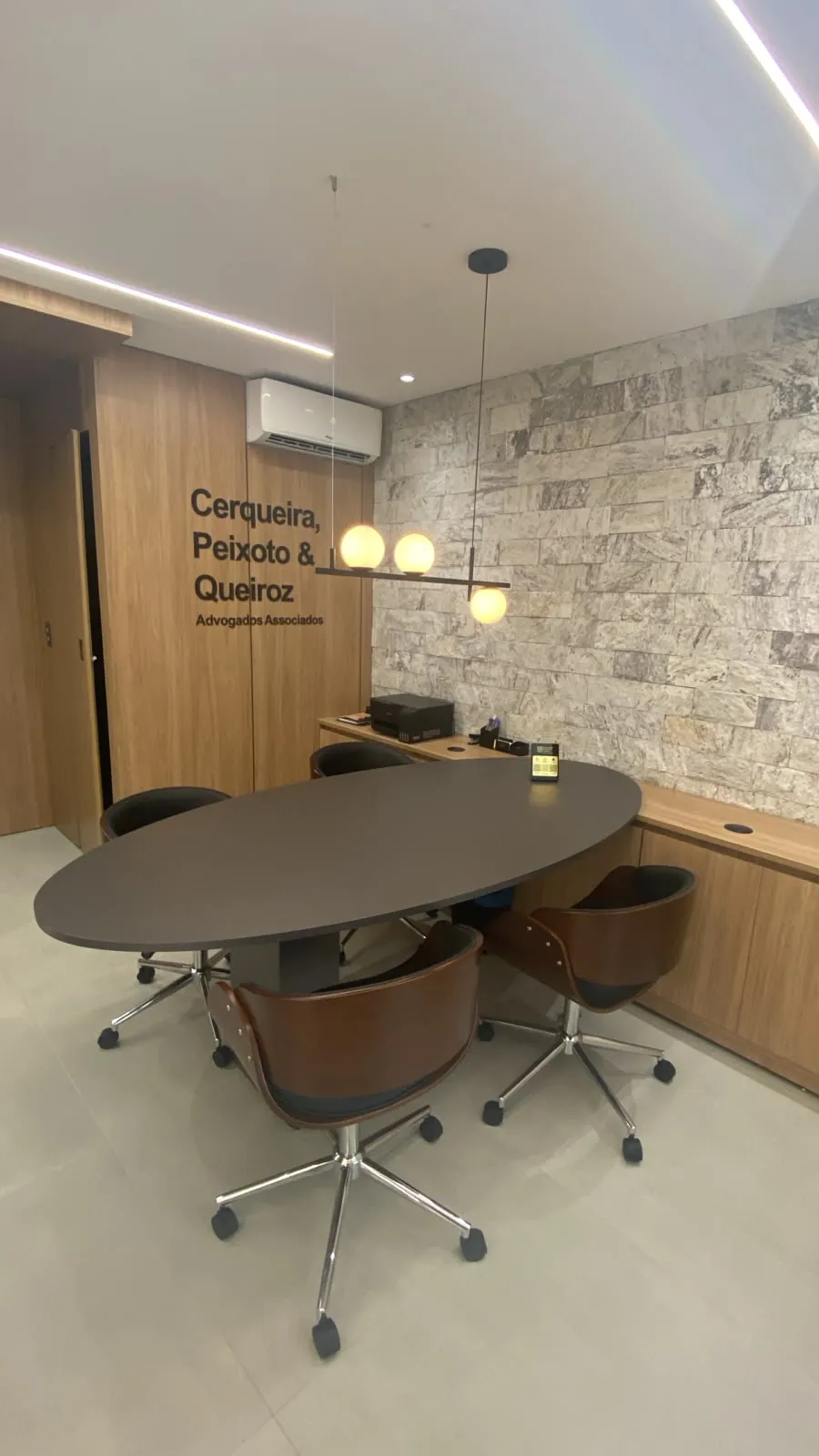 Sala de reunião com mesa oval escura, cadeiras de madeira, parede de pedra e o nome do escritório em letras na parede de madeira.